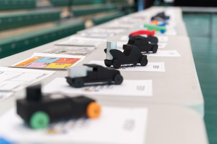 A row of small, student‑designed model race cars displayed on a table, each placed on top of design sheets in a gym setting.